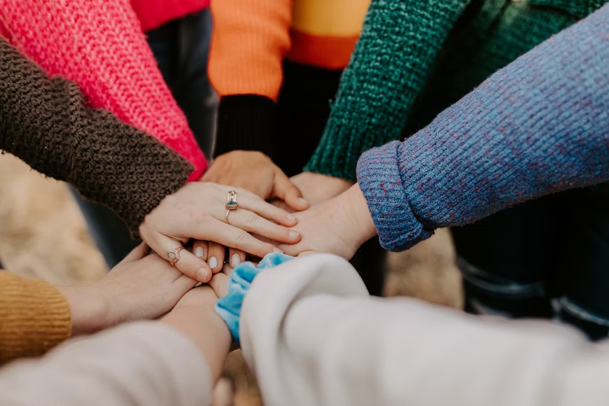 Friends hands piled together, all wearing knitted jumpers and cardigans and the top hand wears three jewelled rings.